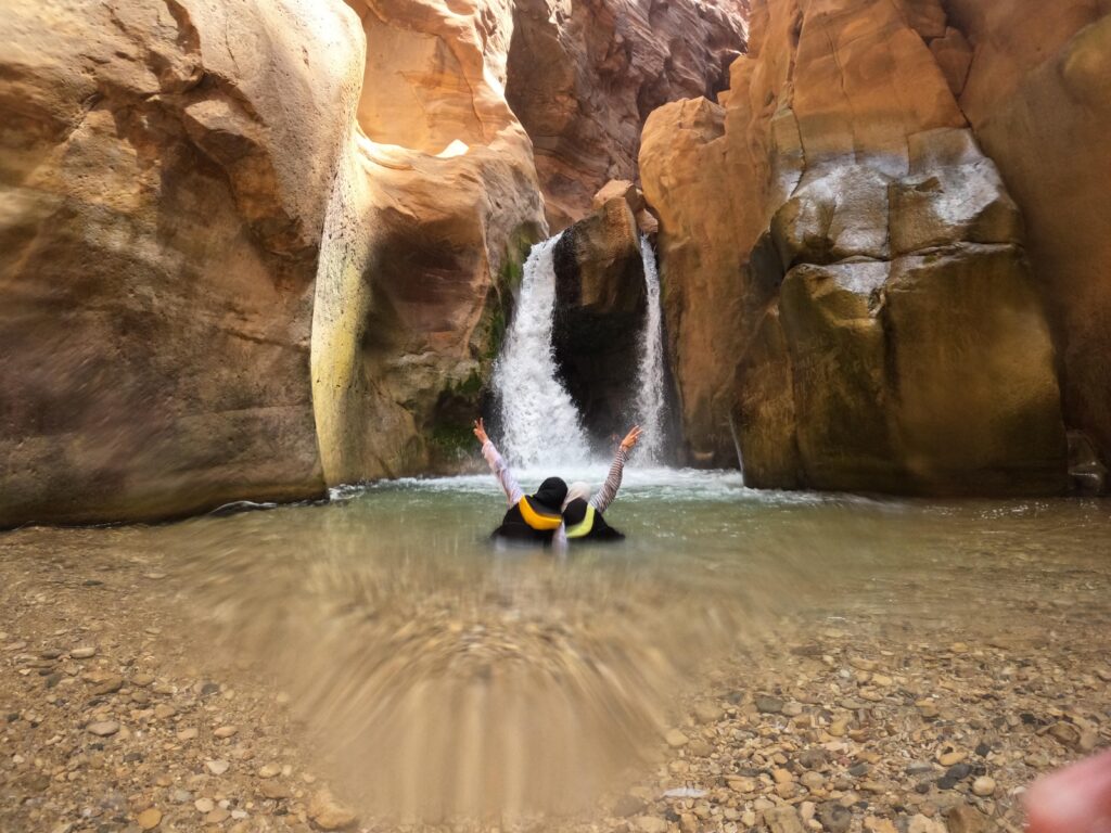 Two people holding each other with the other arm outstretched in front of a waterfall in the Wadi Mujib canyon